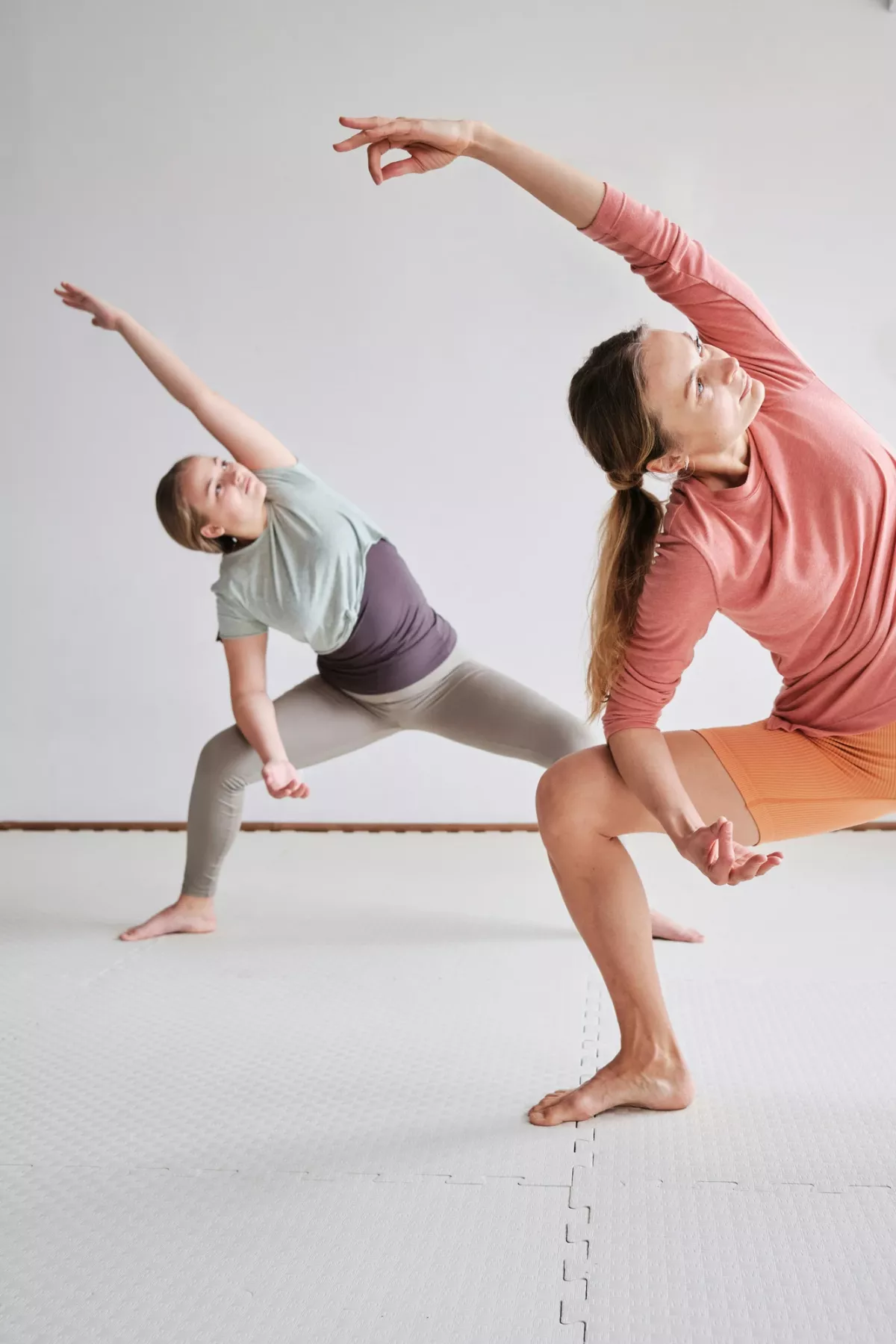 Zwei Frauen praktizieren gemeinsam Yoga in einer Seitendehnung auf wei&szlig;em Boden.