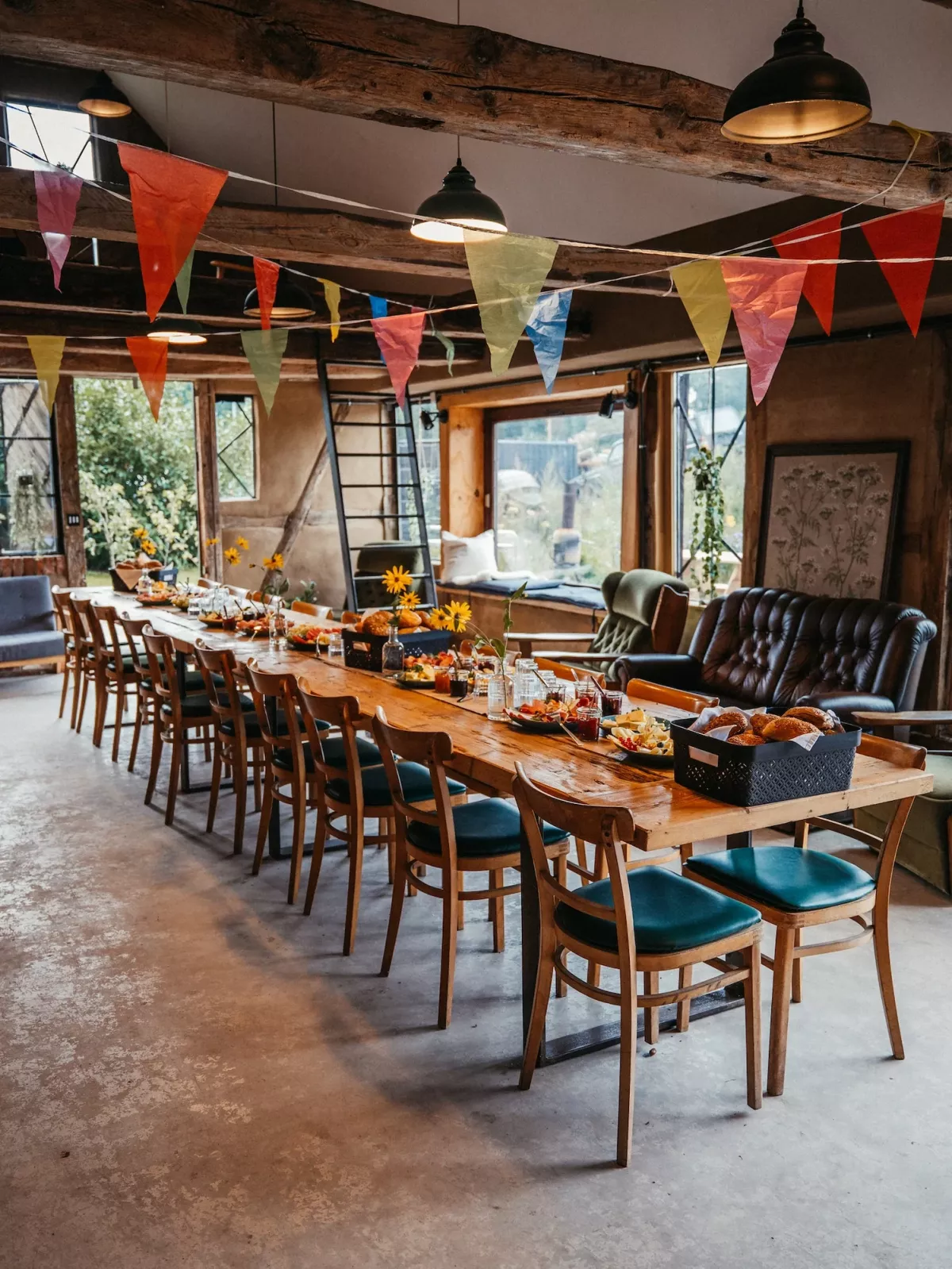 Gro&szlig;er Holztisch mit St&uuml;hlen in festlich dekoriertem Speiseraum mit bunten Girlanden und schwarzen Pendelleuchten f&uuml;r Teambuilding-Events.