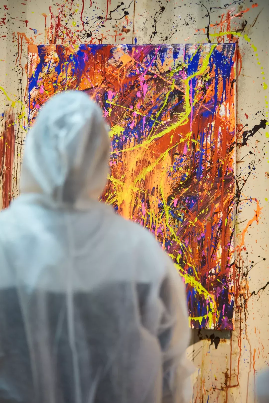 Person von hinten in wei&szlig;em Schutzanzug betrachtet buntes abstraktes Gem&auml;lde mit roten, gelben und blauen Farbspritzern an einer Wand.
