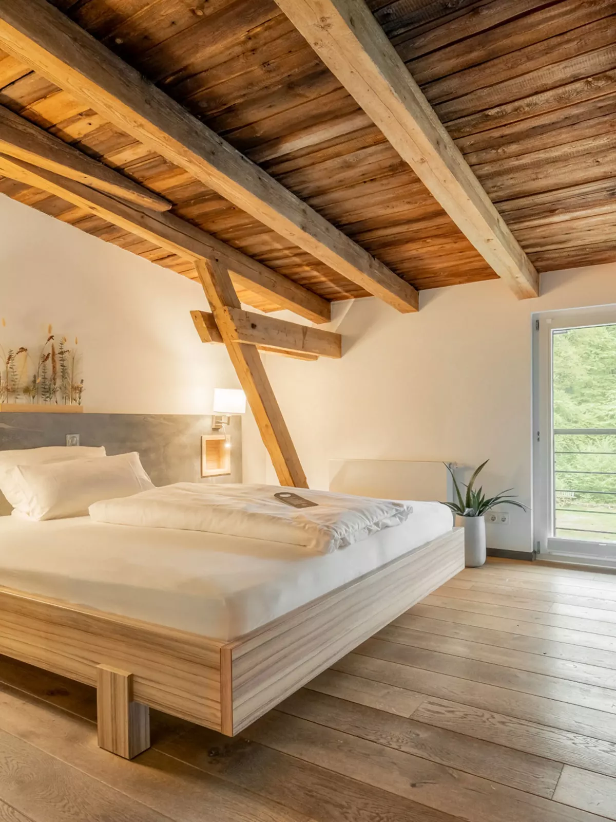 Helles Dachzimmer mit Holzbalkendecke, Doppelbett aus Holz, Nachttischen mit Lampen und gro&szlig;en Fenstern mit Ausblick ins Gr&uuml;ne.