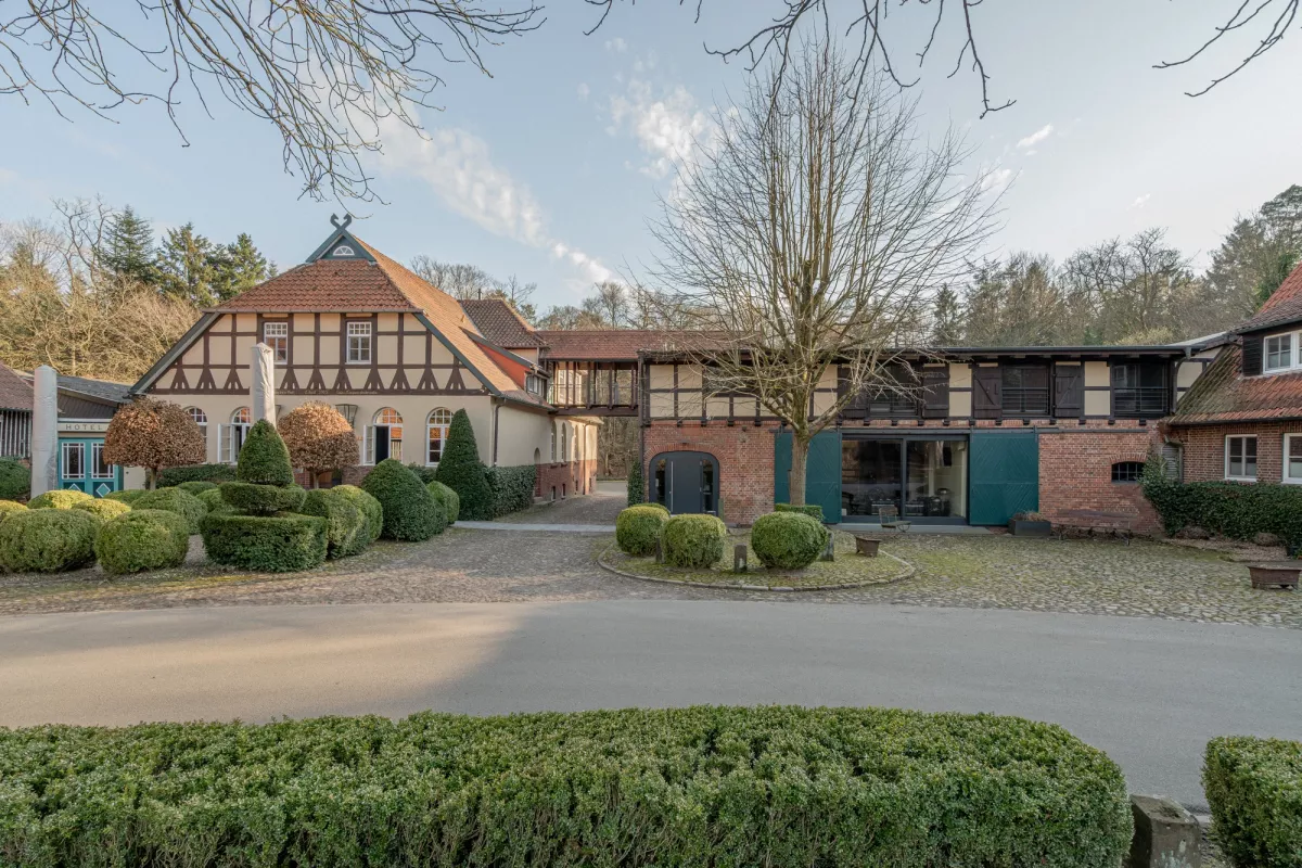 Historisches Fachwerkhaus mit Ziegeldach und gepflegtem Vorgarten, umgeben von B&auml;umen und gr&uuml;nen Hecken.