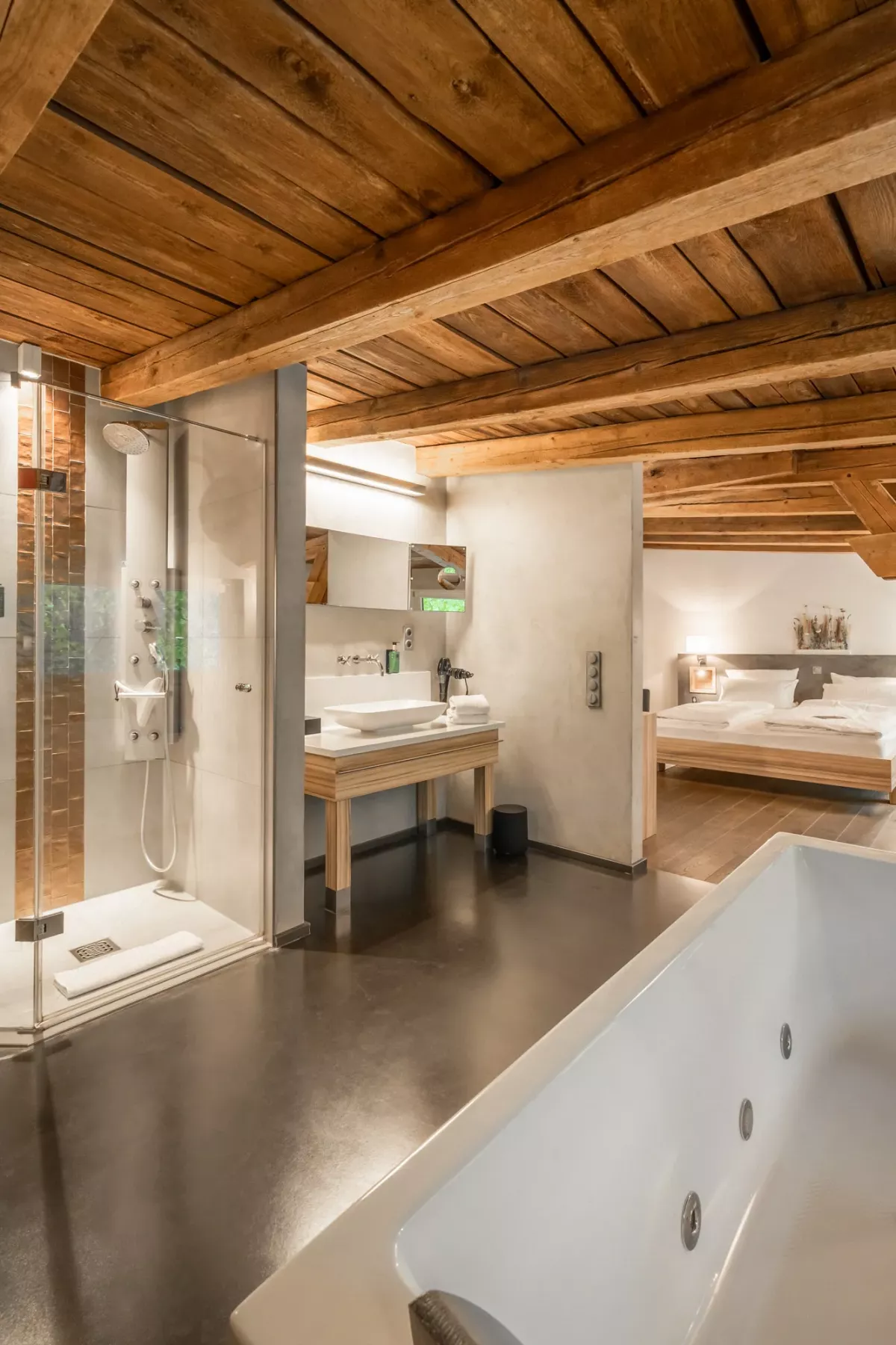 Luxuri&ouml;ses Badezimmer mit Holzbalkendecke, Whirlpool, begehbarer Dusche und Doppelwaschbecken in heller, moderner Gestaltung. Schlafbereich im Hintergrund.