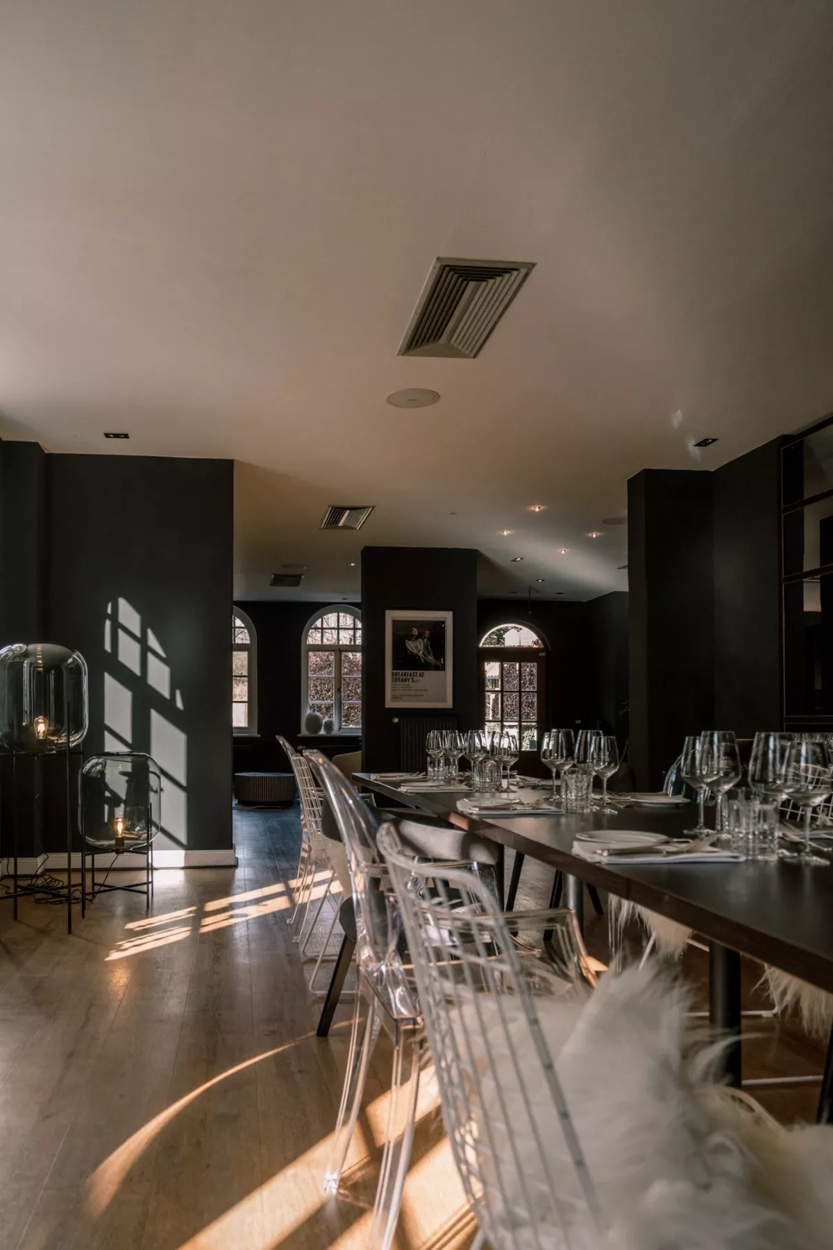 Eleganter Veranstaltungsraum mit dunklen W&auml;nden, langer Holztafel und gepolsterten St&uuml;hlen, Sonnenlicht f&auml;llt durch gro&szlig;e Fenster.