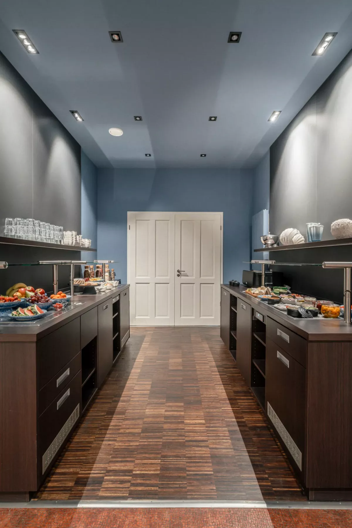 Moderne Buffetzone mit blauen W&auml;nden, dunklen Holzarbeitsplatten, Ausstellungstheken mit Speisen und wei&szlig;er T&uuml;r im Hintergrund.