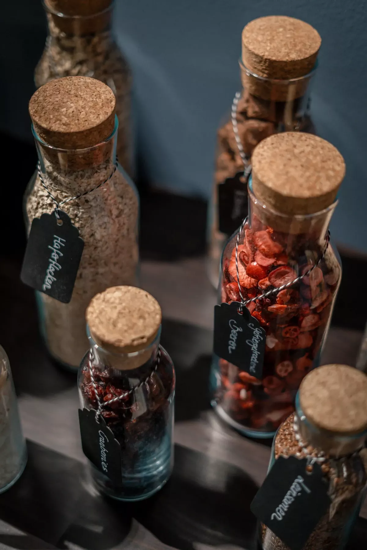 Glasflaschen mit Korkstopfen gef&uuml;llt mit Haferflocken, getrockneten Erdbeeren, getrockneten Himbeeren und M&uuml;sli, arrangiert auf dunkler Oberfl&auml;che.