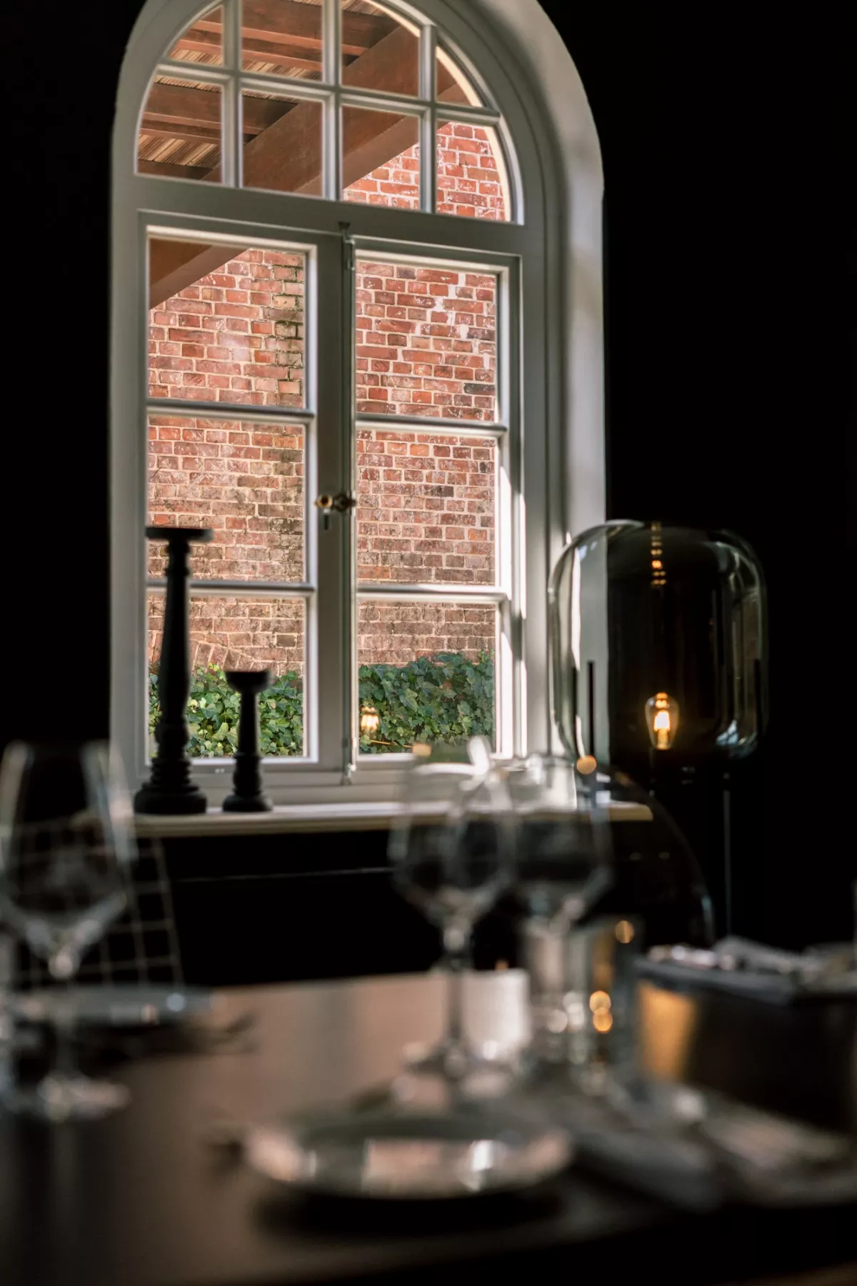 Elegantes Speisezimmer mit gro&szlig;em Bogenfenster auf Backsteinwand, Weingl&auml;sern und Kerzenleuchtern im Vordergrund.