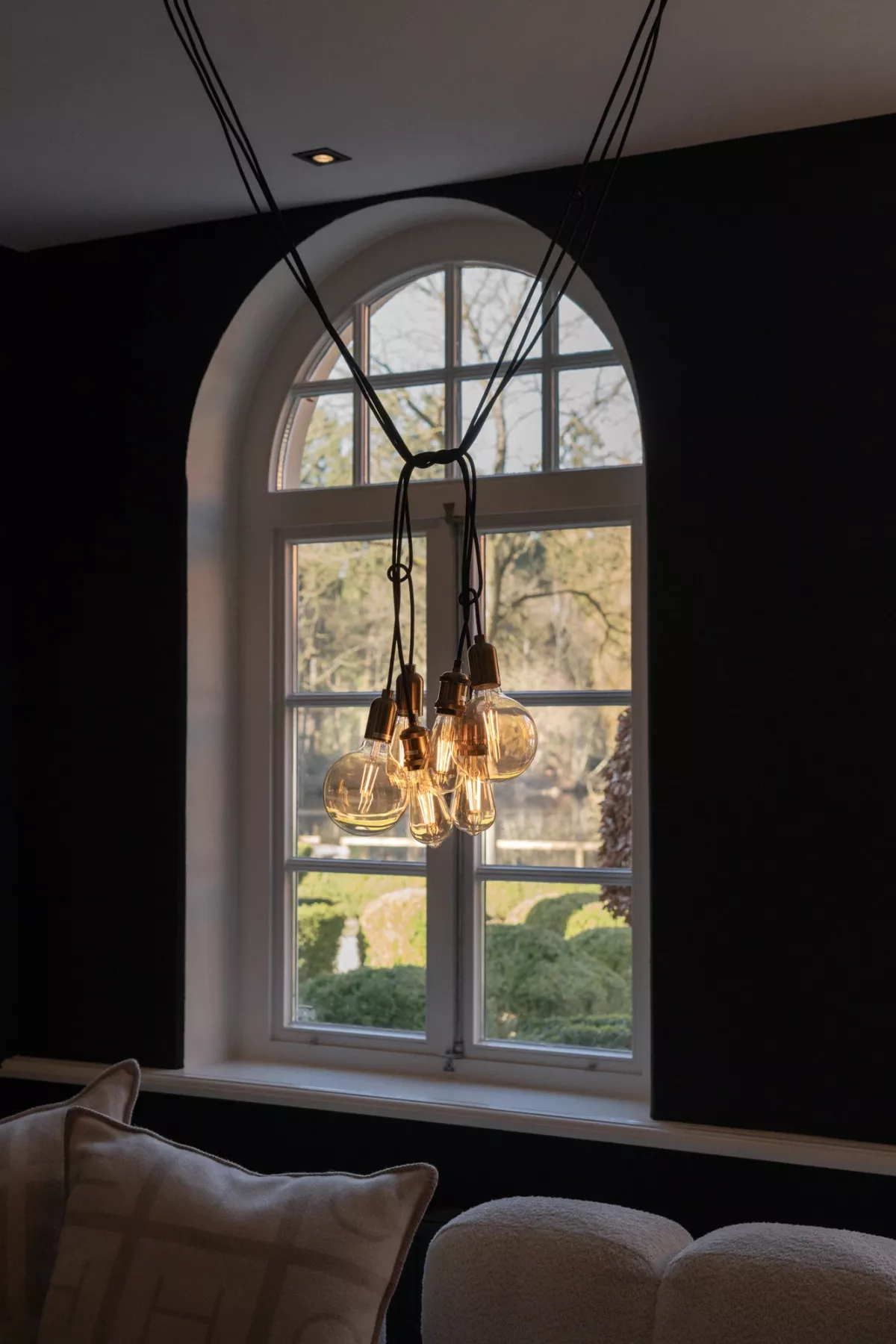 Gro&szlig;es Rundbogenfenster mit Ausblick auf gr&uuml;ne Landschaft, darunter h&auml;ngt eine moderne Pendelleuchte aus Vintage-Gl&uuml;hbirnen.
