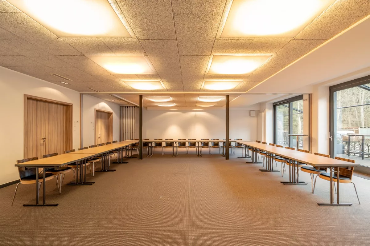 Gro&szlig;er heller Veranstaltungsraum mit Holzdecke, Reihen von Tischen und St&uuml;hlen, gro&szlig;en Fenstern und modernen Deckenleuchten.