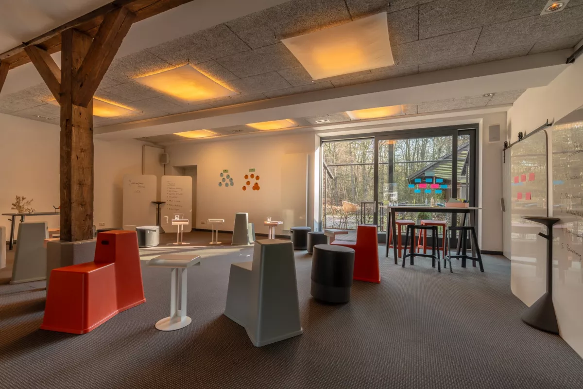 Moderner Tagungs- und Workshopraum mit Holzbalken, beleuchteten Deckenfeldern, orangenen Sitzelementen und gro&szlig;en Fenstern mit Blick ins Gr&uuml;ne.