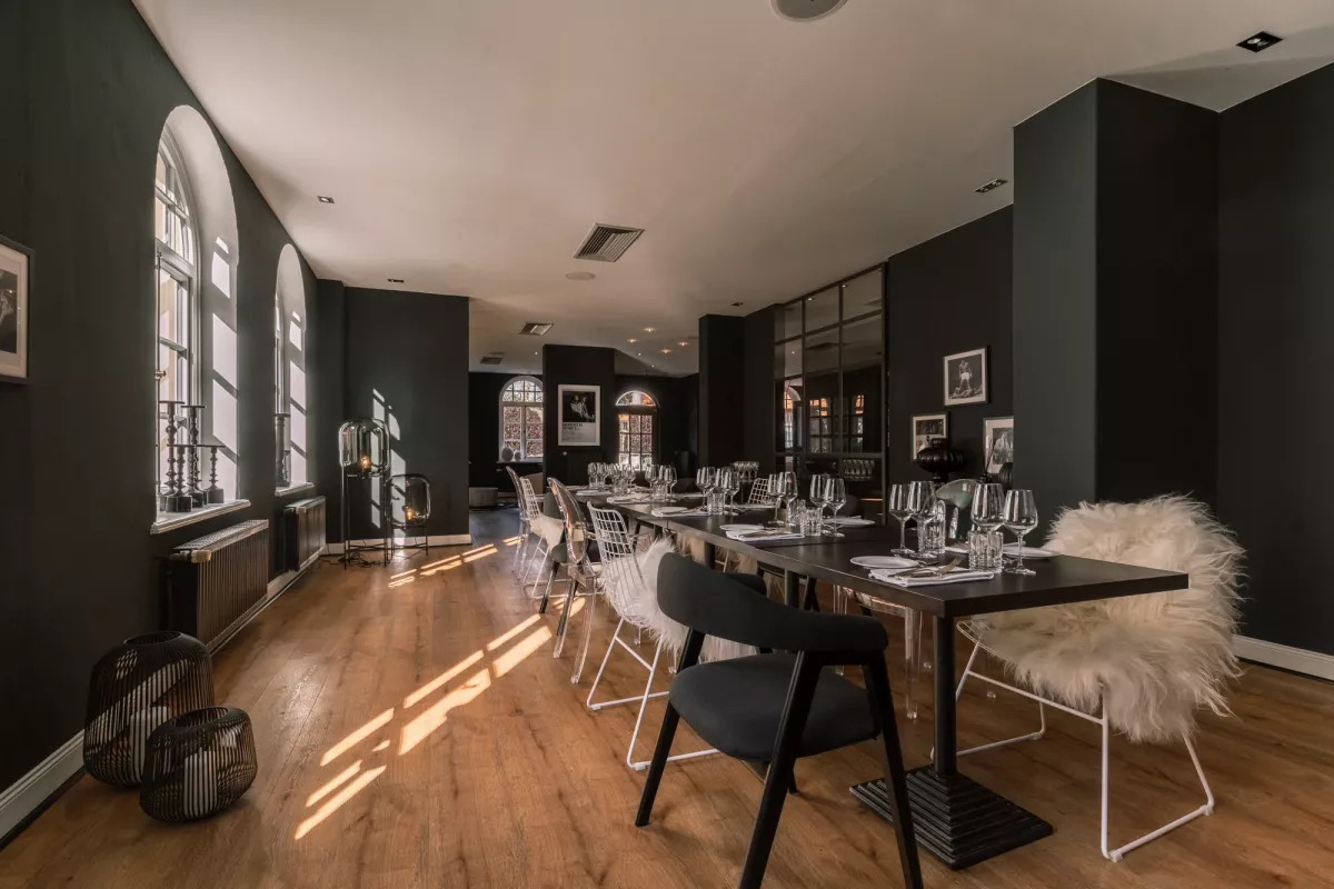 Eleganter Veranstaltungsraum mit schwarzen W&auml;nden, Holzboden und gro&szlig;en Fenstern. Eine lange Tafel mit wei&szlig;en St&uuml;hlen und Fell-Dekoration.