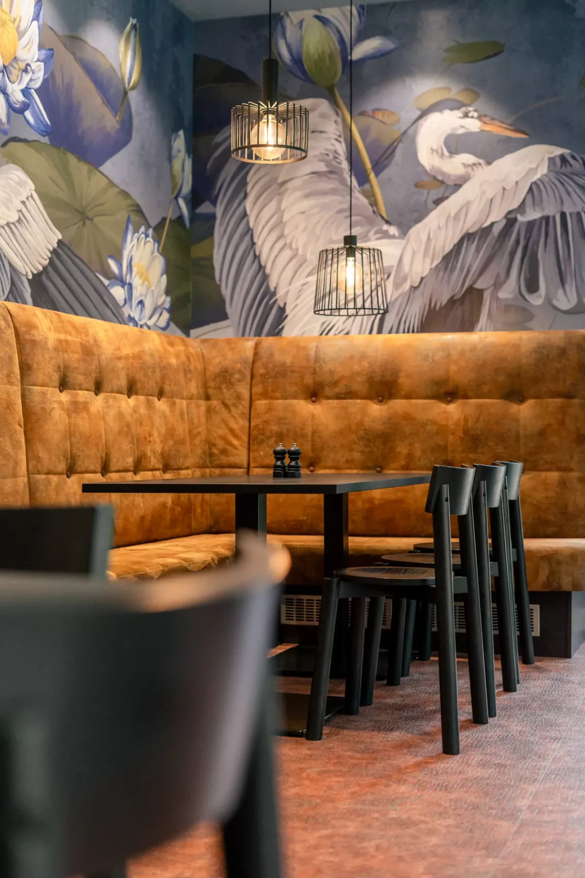 Gem&uuml;tliche Restaurantecke mit orangefarbenem Samtsofa, schwarzen St&uuml;hlen und Tisch vor einer Wandmalerei mit wei&szlig;en Reihern und h&auml;ngenden Lampen.