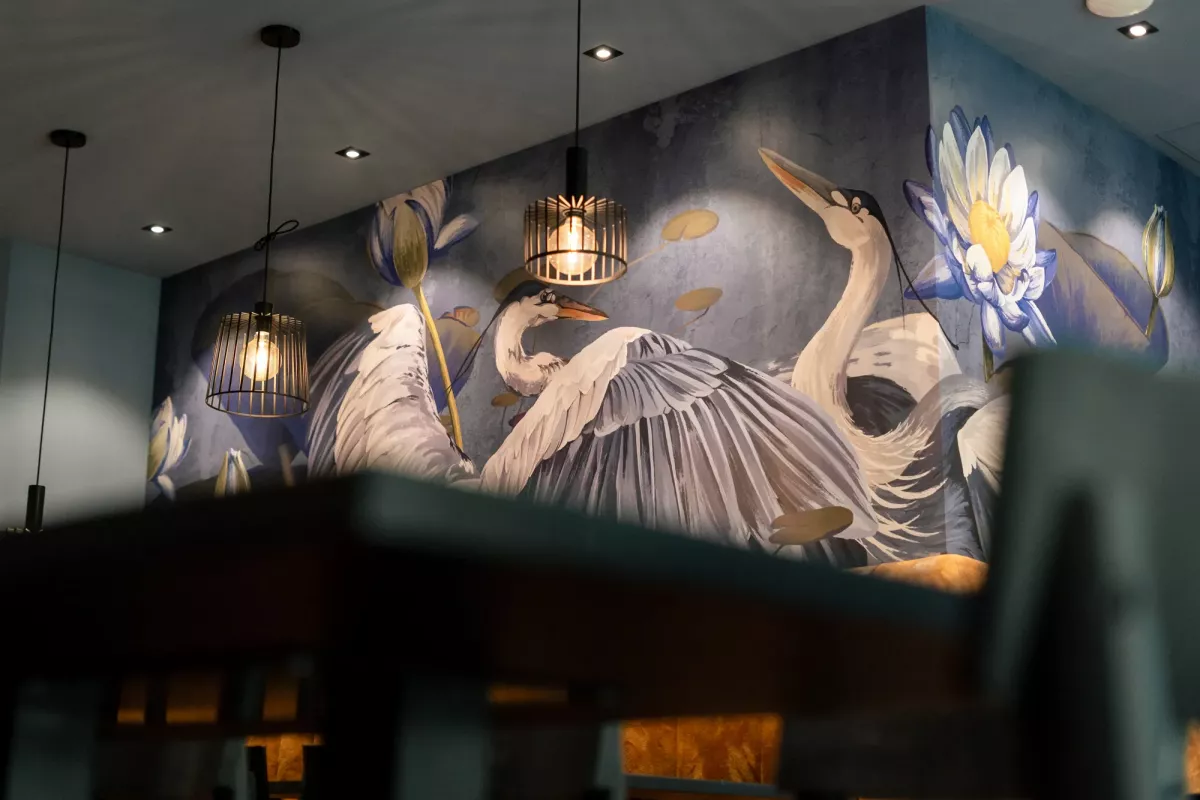 Elegante Wandmalerei mit wei&szlig;en Reihern und Wasserlilien an dunkler Wand, illuminiert durch h&auml;ngende Lampen in gehobener Eventlocation.
