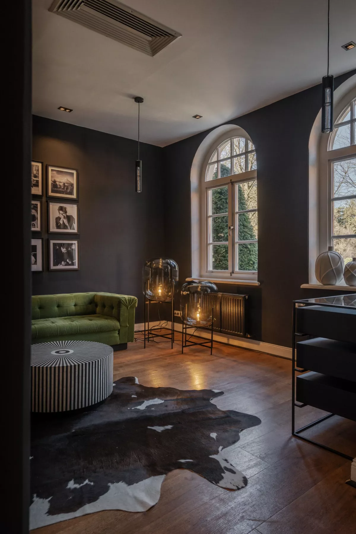Elegante Lounge mit dunkelgrauen W&auml;nden, gr&uuml;nem Samt-Sofa, gro&szlig;em Rundbogenfenster, Holzboden und schwarzwei&szlig;er Kuhfellmatte.