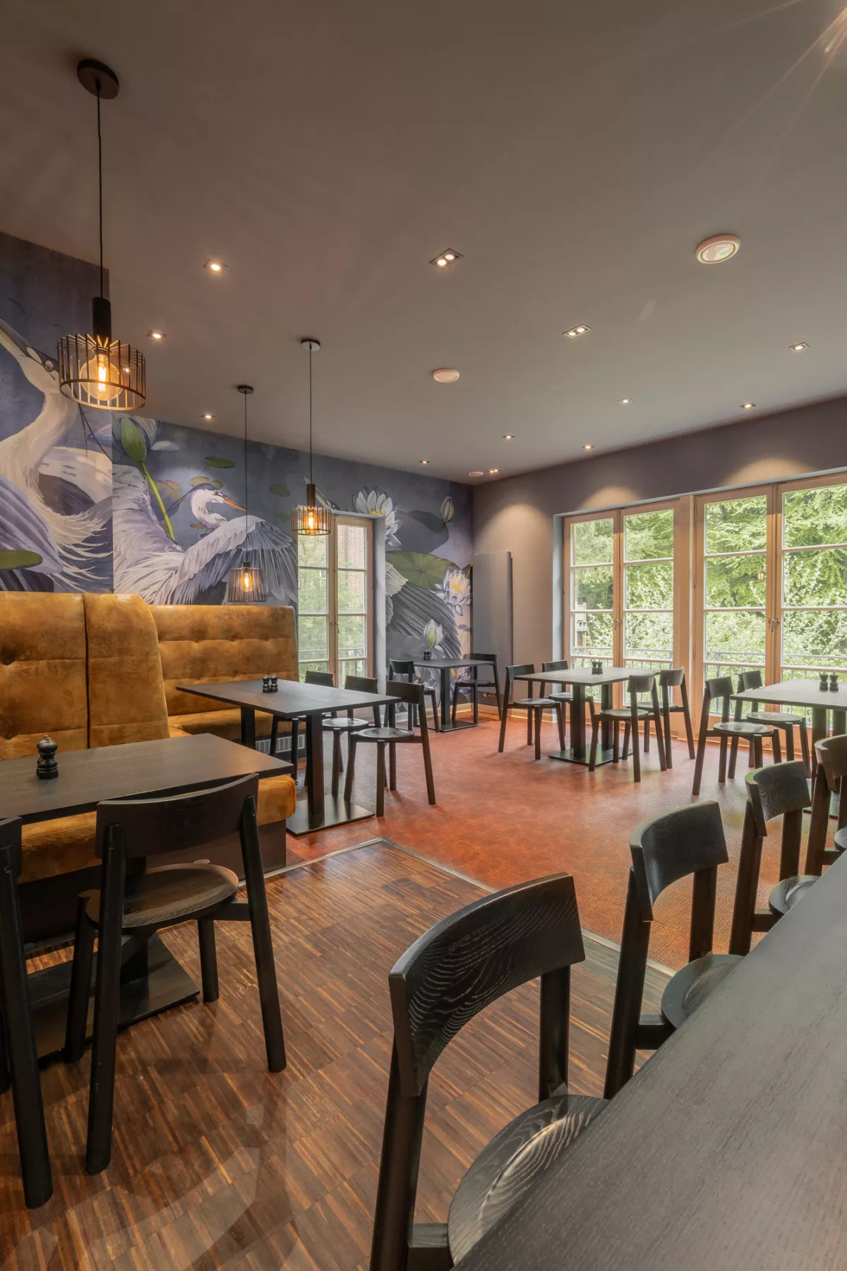 Moderner Fr&uuml;hst&uuml;cksraum mit cognacfarbenen Polsterm&ouml;beln, dunklen Holztischen, gro&szlig;fl&auml;chigen Wandbildern und Blick auf Wald.